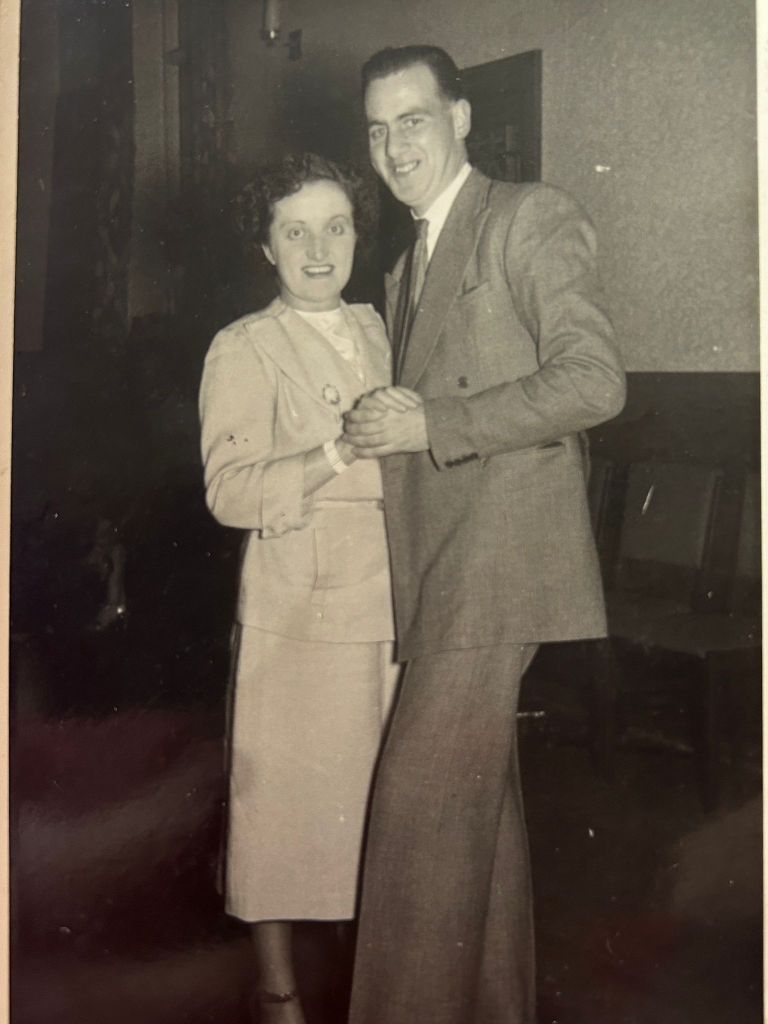 Black-and-white photograph of Rita and Denis, an Irish diasporic couple who met in Birmingham, standing close together and smiling at a Birmingham ballroom. Rita, much shorter than Denis, wears a light skirt suit and has short dark curly hair. Denis wears a mid-grey suit and tie and has short, slicked back dark hair. They are holding hands as if dancing.